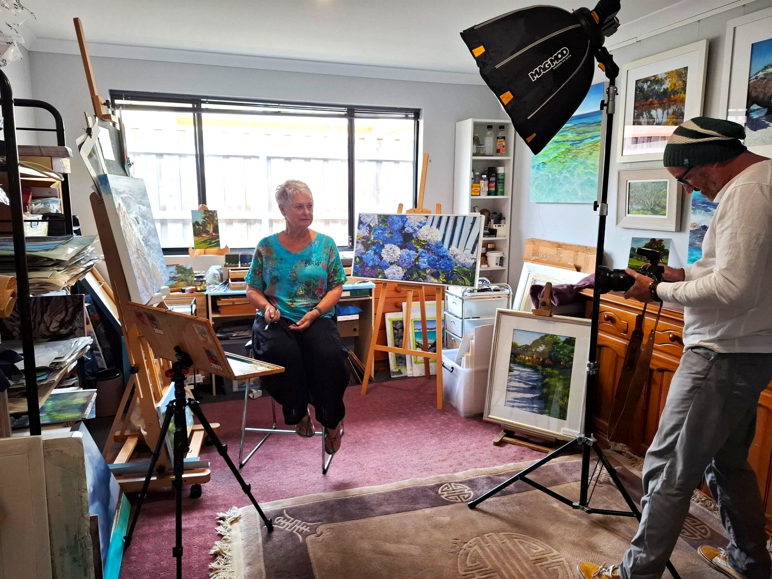 A woman sits in her art studio surrounded by paintings, while a man holding a camera takes pictures of her